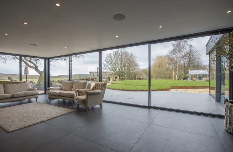 A modern living room with large floor-to-ceiling sliding doors opens to a patio, lawn, trees, and a greenhouse outside. Inside, beige sofas, an armchair, and a rug sit atop the tiled floor.