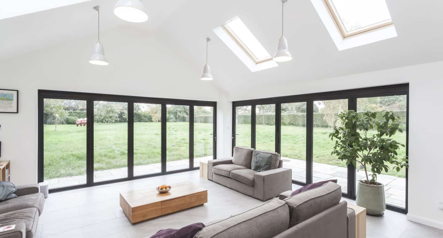 A bright, modern living room with large windows, skylights, and bifold doors opens to a green lawn. Gray sofas, a wooden coffee table, and a potted plant create comfort, while white walls and pendant lights enhance the airy atmosphere.