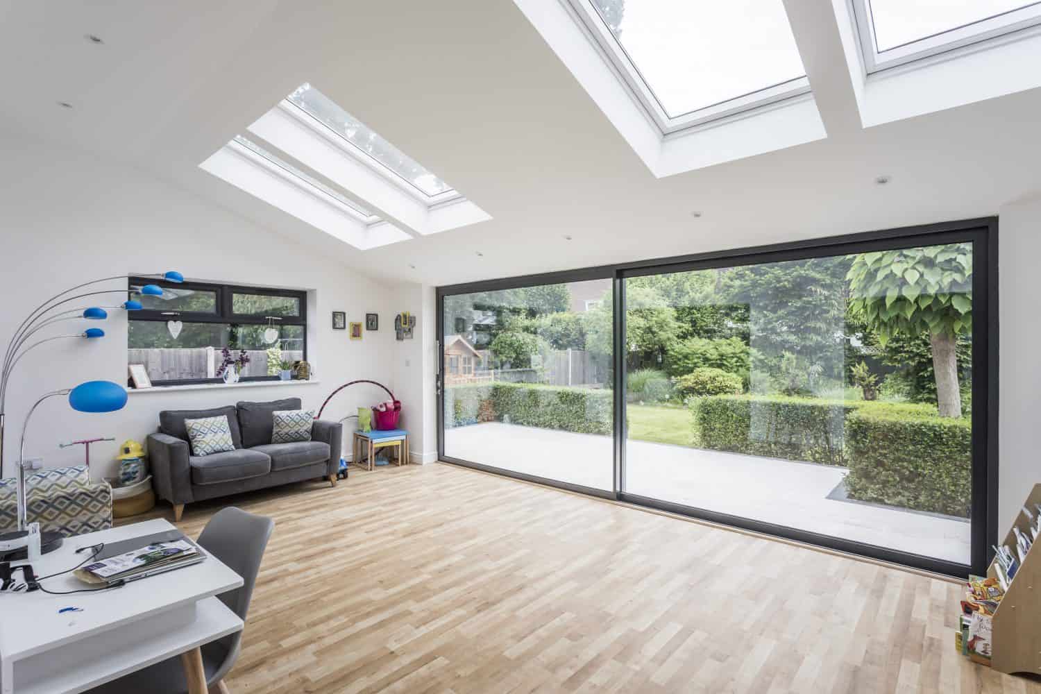 Bright, modern room with wood flooring, skylights, large bifold doors opening to a green garden, a gray sofa, colorful floor lamp, and desk with chair in the corner.