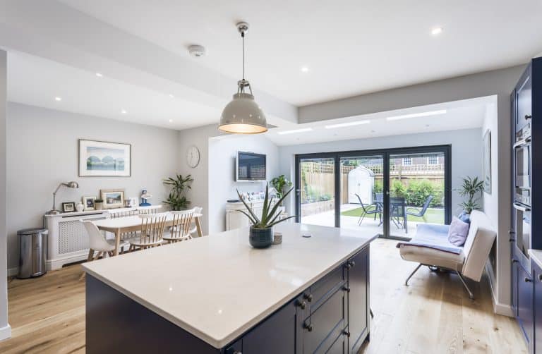 Modern open-plan kitchen and dining area with a large island, wooden floors, stylish decor, and bespoke glass solutions—such as sliding doors—leading to a patio and garden. Natural light fills the bright, spacious room.