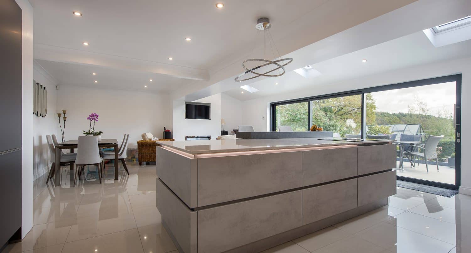 Modern open-plan kitchen and dining area with sleek, light gray cabinetry, glossy tiled floor, bifold doors opening to a patio, and contemporary furnishings in a bright, airy space.
