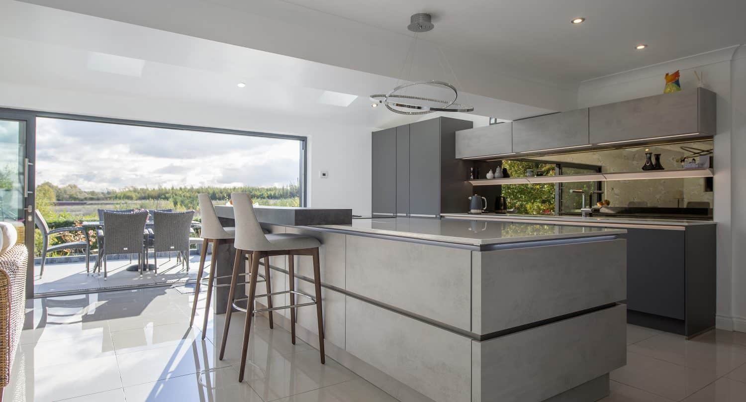 Modern kitchen with sleek grey cabinets, a large island with two bar stools, glossy tiled floor, and sliding doors opening to a patio with outdoor dining furniture and a scenic garden view.