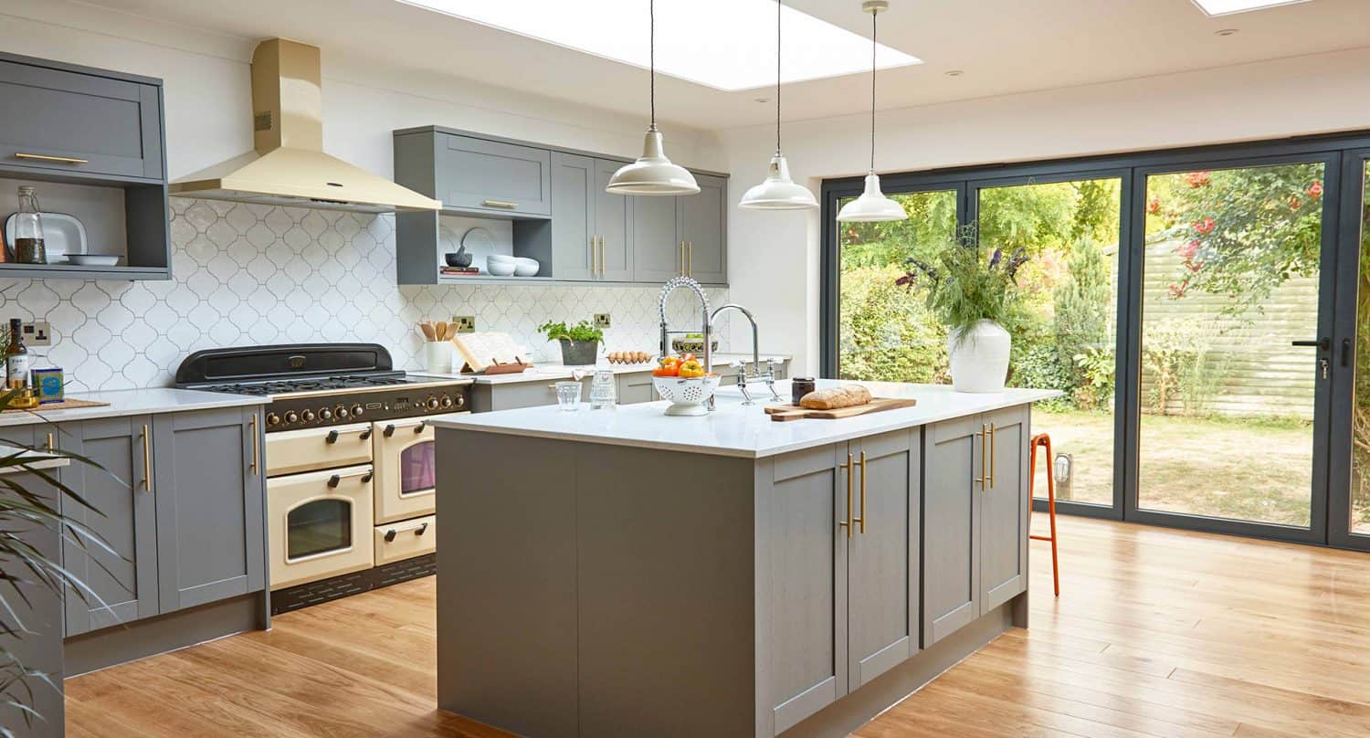 Modern kitchen with grey cabinets, a large island, pendant lights, cream-colored stove, wooden floor, and sliding glass doors opening to a green garden. Glass roofing fills the space with natural light over decor-filled countertops.