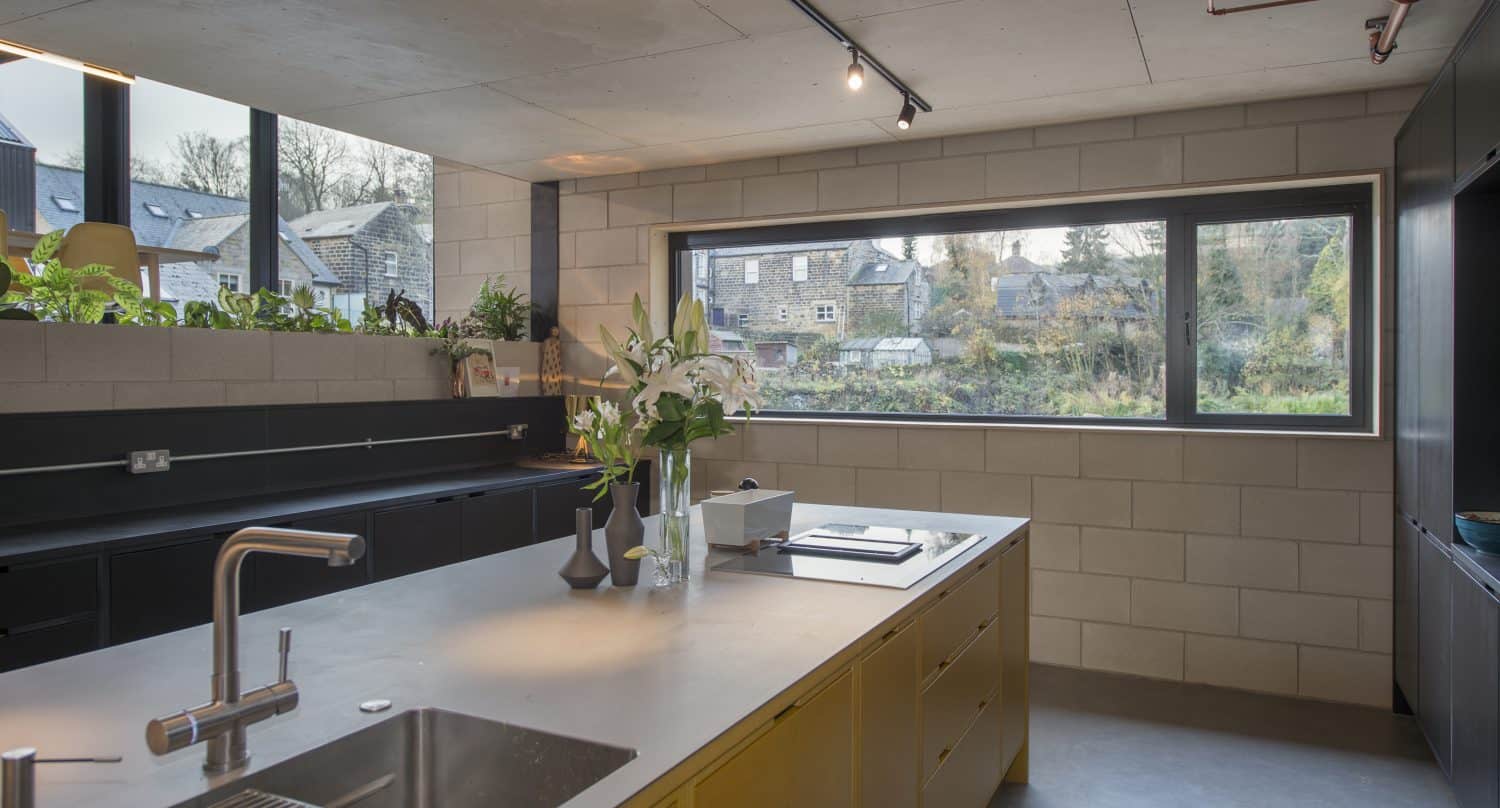 Modern kitchen with a large island, built-in sink, and vases with flowers. Wide bifold doors open to views of houses and greenery. Room features concrete walls, exposed pipes, minimalist decor, and plenty of natural light.