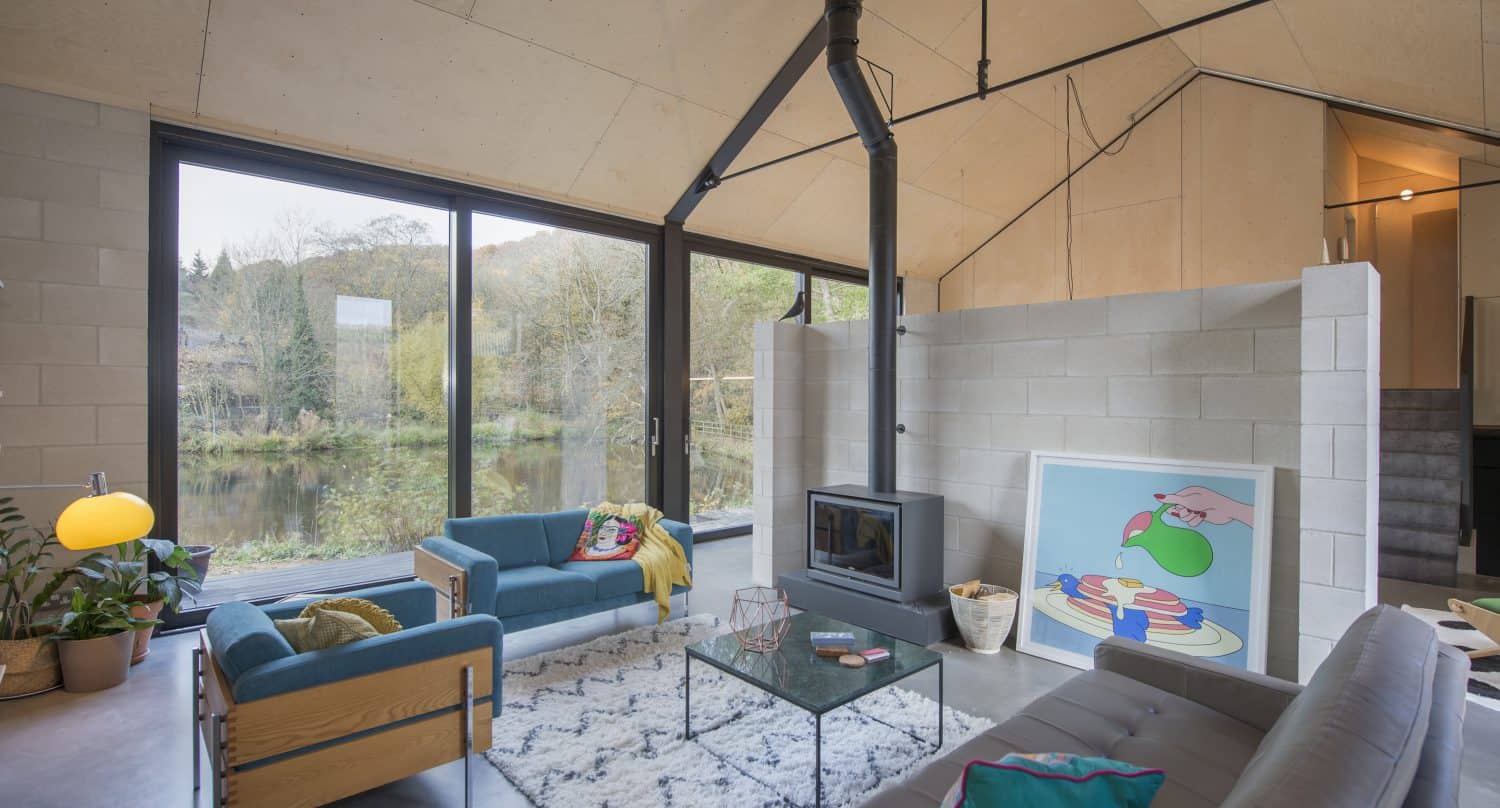 A modern living room with large windows and sliding doors overlooking trees and water. It features blue and gray sofas, a white rug, a wood-burning stove, indoor plants, and colorful artwork against a cinder block wall.