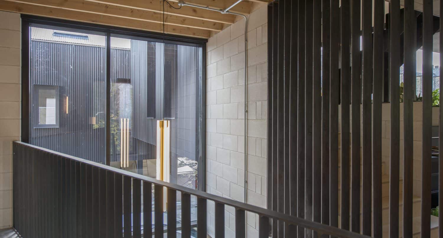 A modern interior hallway with exposed wood beams, concrete block walls, and vertical black metal railings features a large window with bespoke glass solutions, overlooking an outdoor area with similar vertical black paneling. A pendant light hangs near the window.