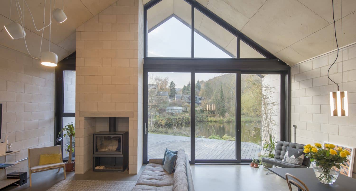 Modern living room with high, slanted glass roofing, large triangular windows, a cozy gray sofa, wood-burning stove, dining table with sunflowers, and views of trees and water outside.