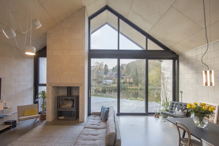 Modern living room with high, slanted glass roofing, large triangular windows, a cozy gray sofa, wood-burning stove, dining table with sunflowers, and views of trees and water outside.