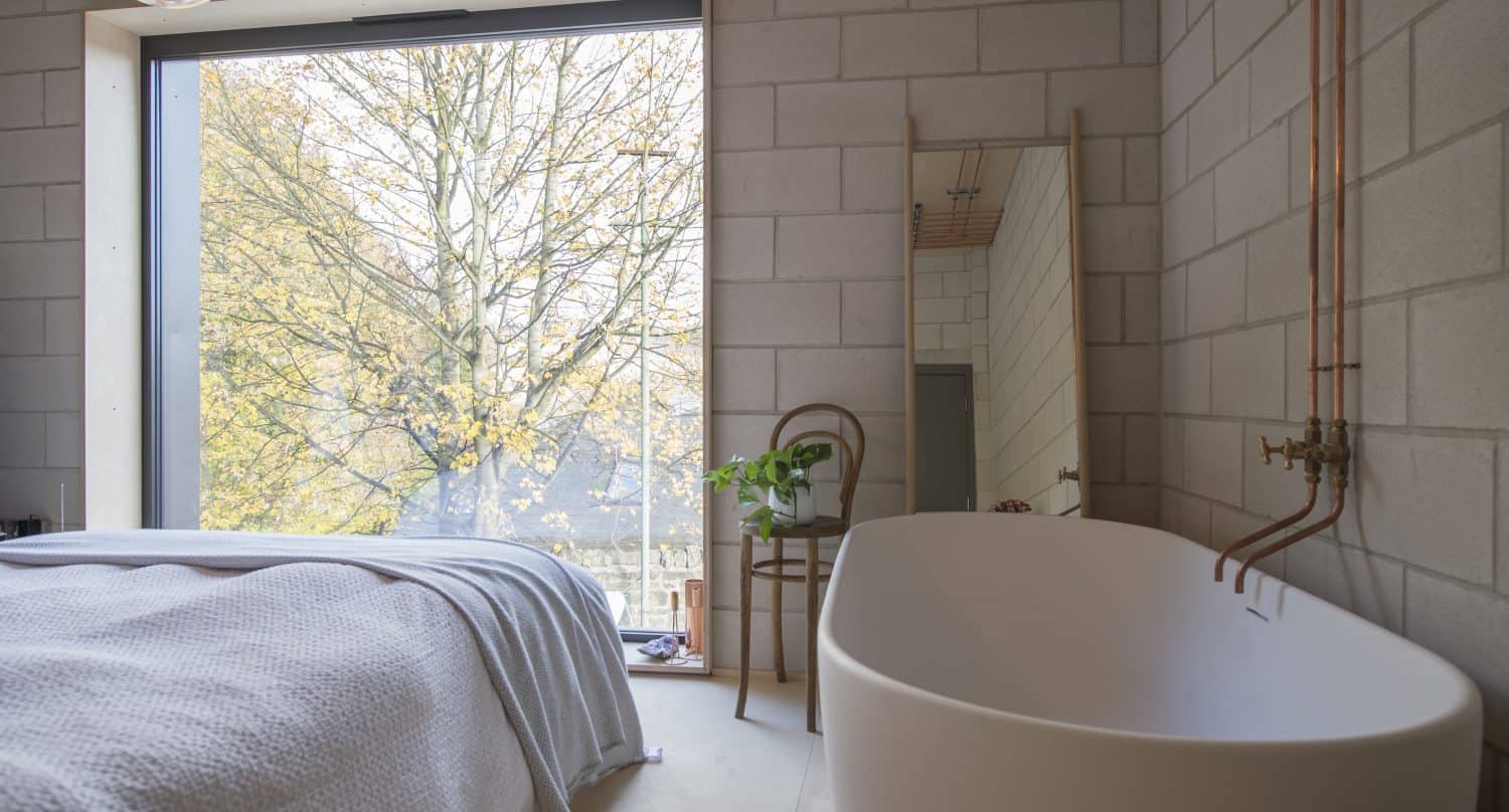 A modern bathroom with a freestanding white bathtub, exposed copper pipes, a wooden chair with a plant, a large mirror, and bifold doors near a floor-to-ceiling window overlooking trees.