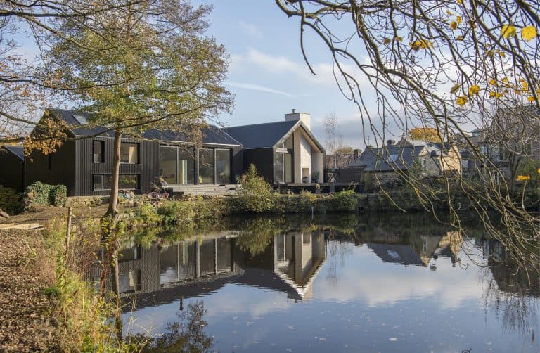 Modern house with large windows and glass roofing sits beside a calm pond, reflecting the building and autumn trees. The sky is clear, and other houses are visible in the background.
