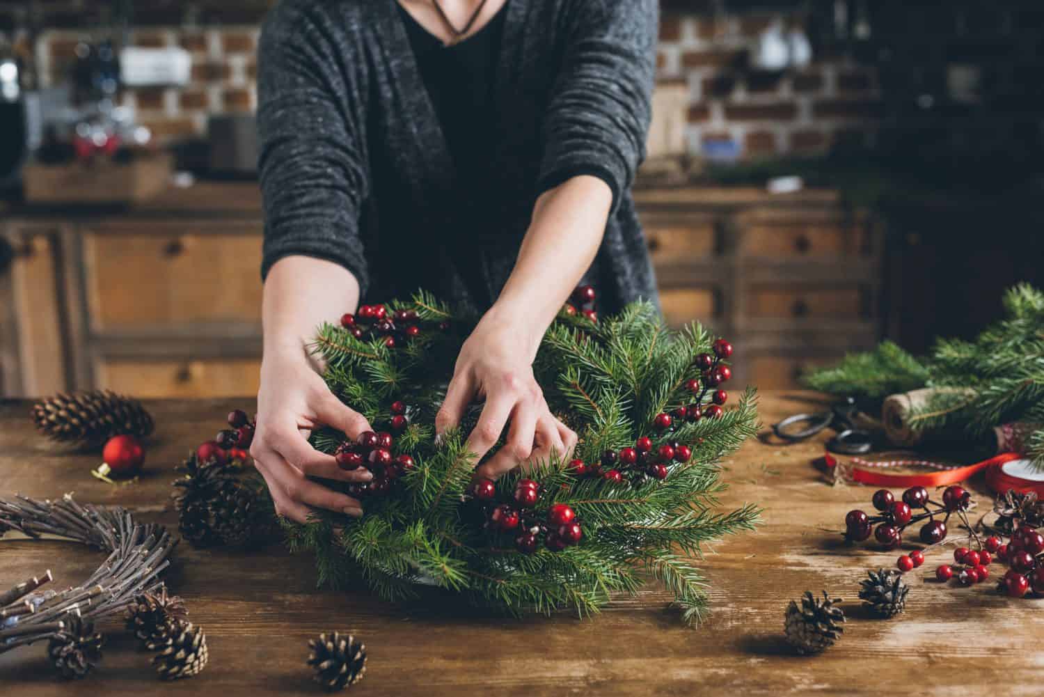 cropped view of florist making Christmas wreath of fir branches, decorative berries and pine cones at workplace