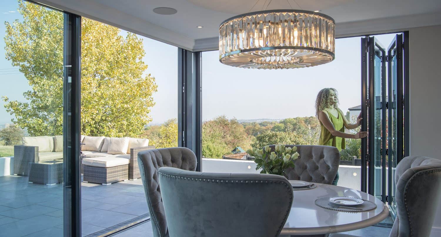 A woman stands by open sliding doors leading from a modern dining area to a spacious outdoor patio with seating, surrounded by trees and a scenic view. The room features elegant chairs and a chandelier.