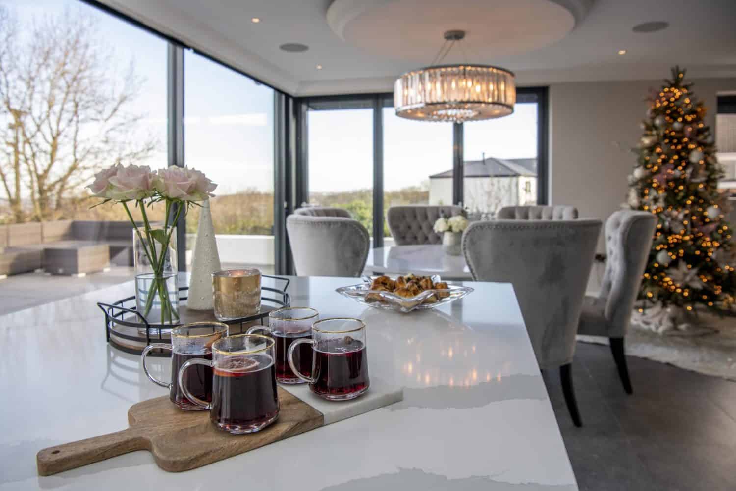 Four glass mugs of mulled wine on a wooden board, a plate of pastries, and a vase of roses sit on a kitchen island in a bright room with bespoke glass solutions, sliding doors, a Christmas tree, and large windows overlooking the patio.