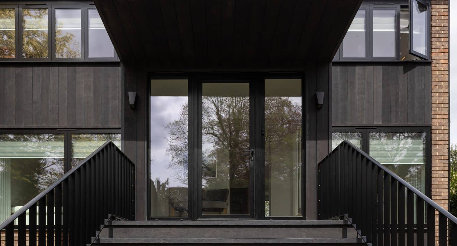 A modern building entrance features wide black steps leading up to bespoke glass solutions in double doors, framed by dark wood paneling and large windows on either side. Trees are reflected in the glass.