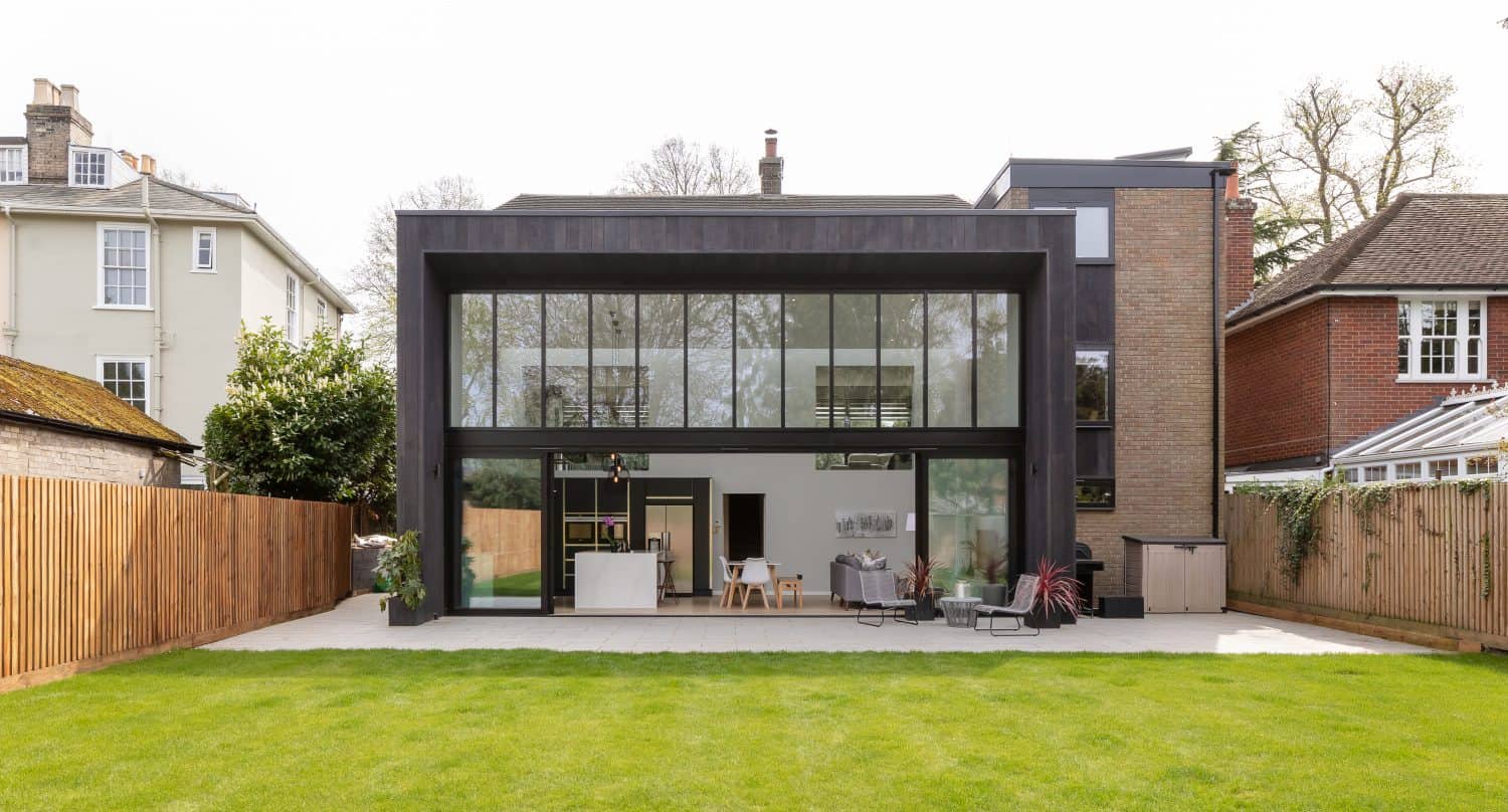 A modern house with large glass windows and a dark exterior, featuring sliding doors that open to an inviting patio area with outdoor seating and a dining table, all surrounded by a green lawn and wooden fences.