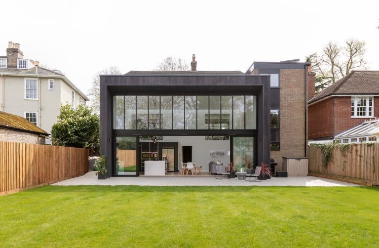 A modern house with large glass windows and a dark exterior, featuring sliding doors that open to an inviting patio area with outdoor seating and a dining table, all surrounded by a green lawn and wooden fences.
