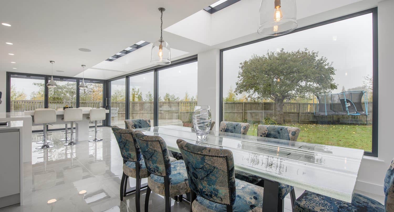 Modern dining area with a glass table and eight blue patterned chairs, large windows and bifold doors opening to a garden with green grass, trees, and a trampoline; white bar stools are visible near the kitchen island.