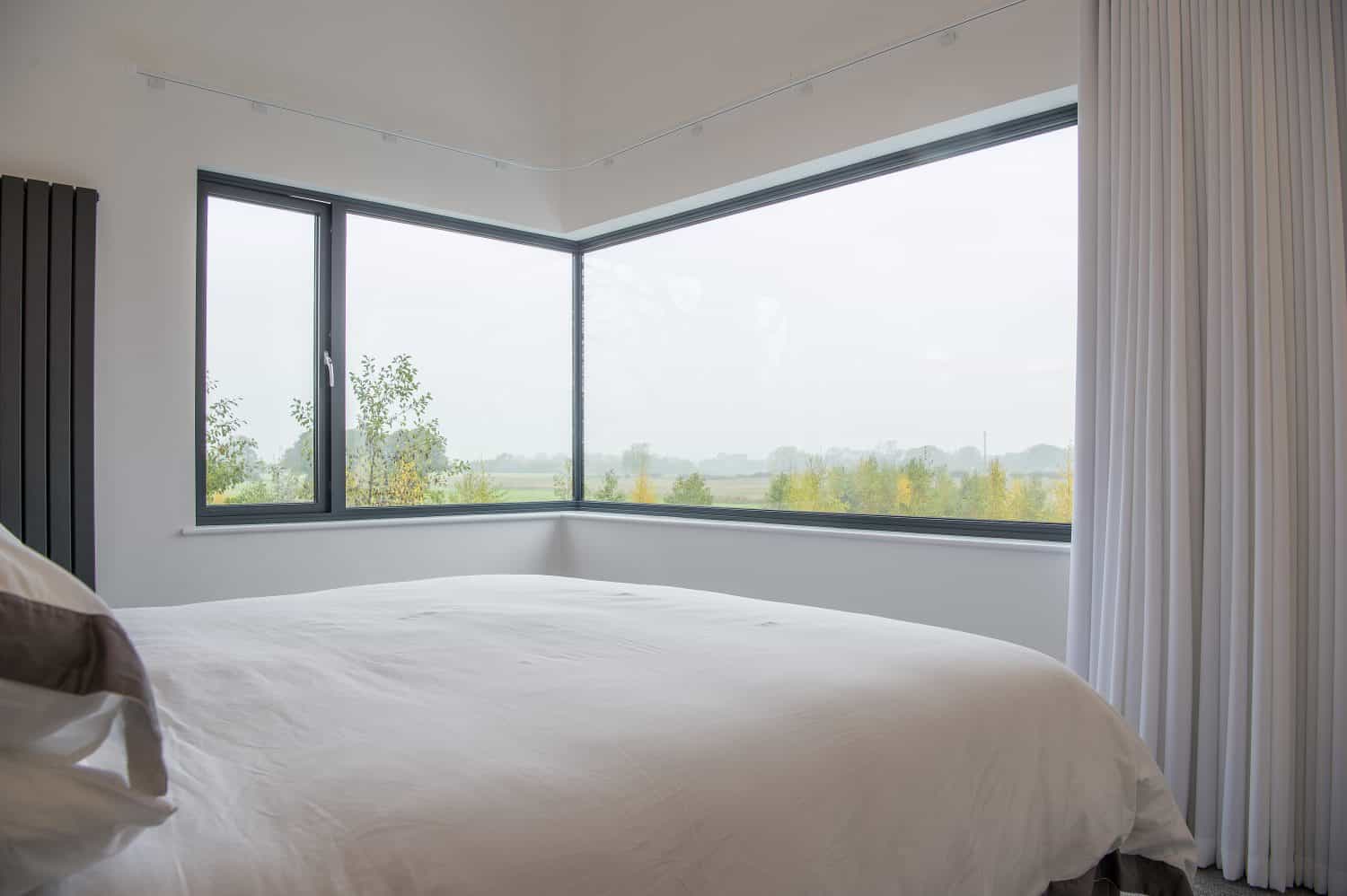 A modern bedroom with a large bed covered in white bedding features bespoke glass solutions in the form of floor-to-ceiling corner windows, offering views of a foggy landscape with trees and fields outside. White curtains partially frame the window.