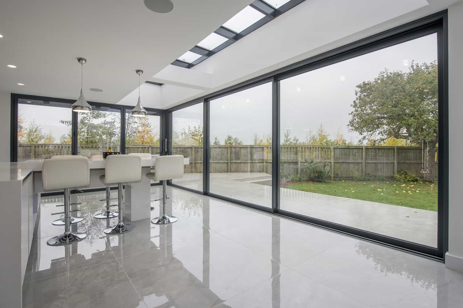 Modern kitchen with white bar stools, pendant lights, and large floor-to-ceiling sliding doors opening to a fenced backyard with trees and a lawn. The room features glossy tiled floors, skylights above, and bespoke glass solutions throughout.