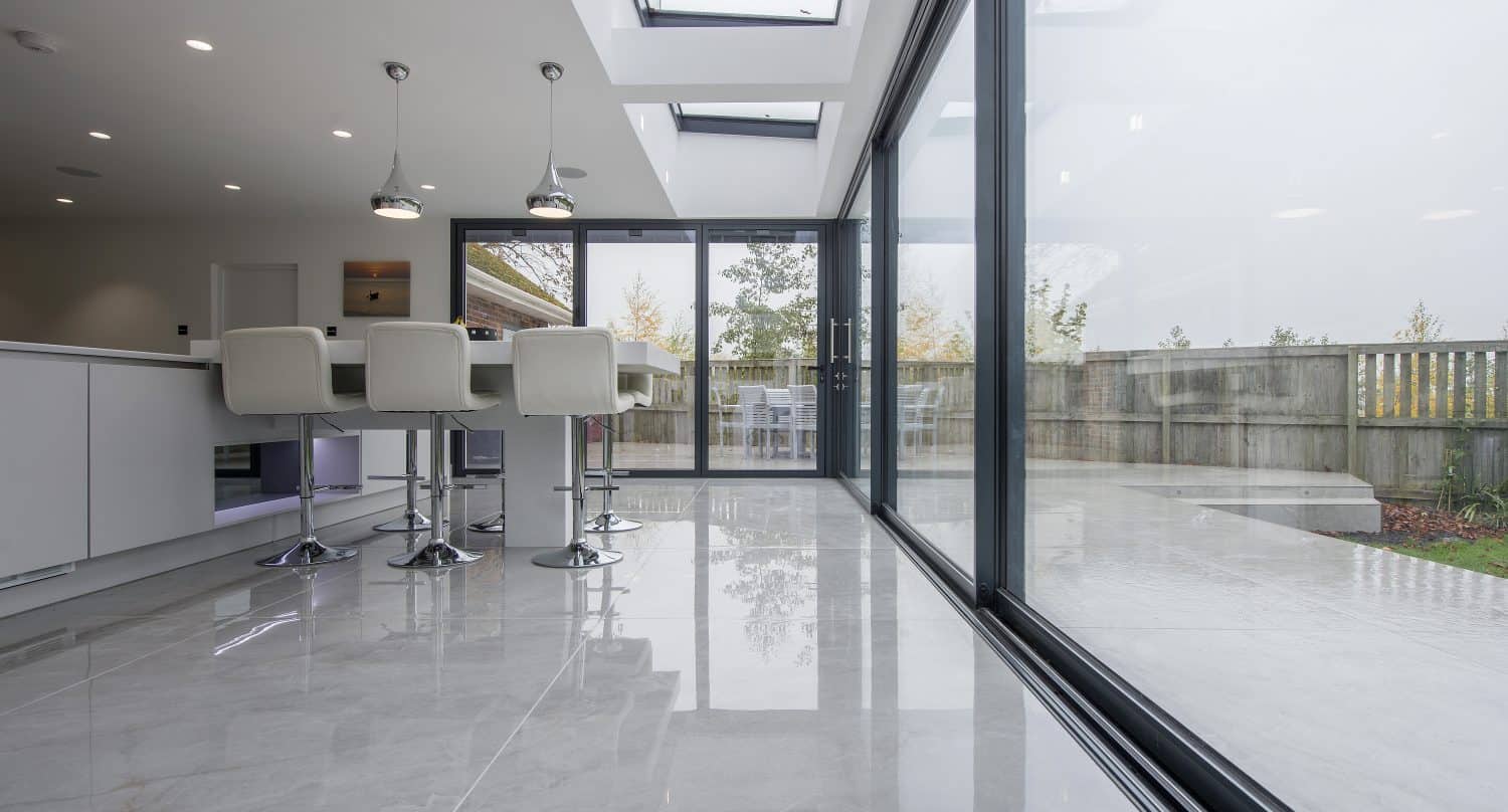 Modern kitchen with glossy tiled floor, white bar stools at an island, pendant lights, and large glass bifold doors opening to a patio with outdoor seating and a fenced yard. Skylights let in plenty of natural light.