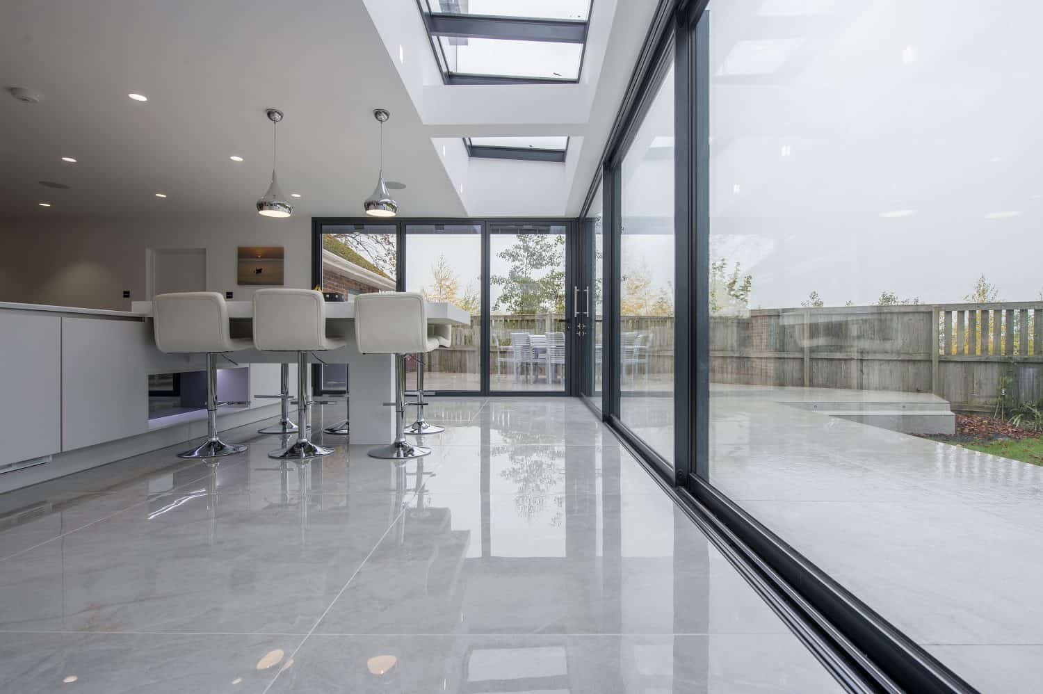 Modern kitchen with glossy tiled floor, white bar stools at an island, pendant lights, and large glass bifold doors opening to a patio with outdoor seating and a fenced yard. Skylights let in plenty of natural light.