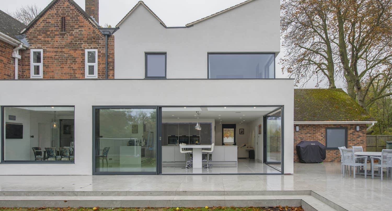 A modern house with a white exterior and bespoke glass solutions, featuring large sliding doors opening onto a spacious patio with outdoor dining furniture, next to a lawn and surrounded by trees.