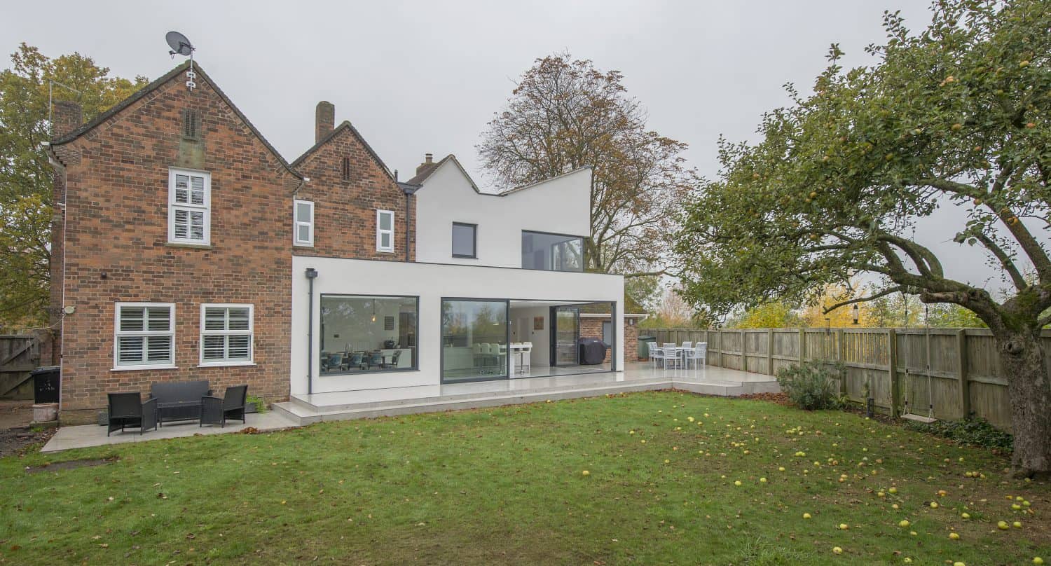 A modern house with a mix of red brick and white exterior walls, large bifold doors, a patio area with outdoor seating, and a spacious grassy backyard with a tree and wooden fence.