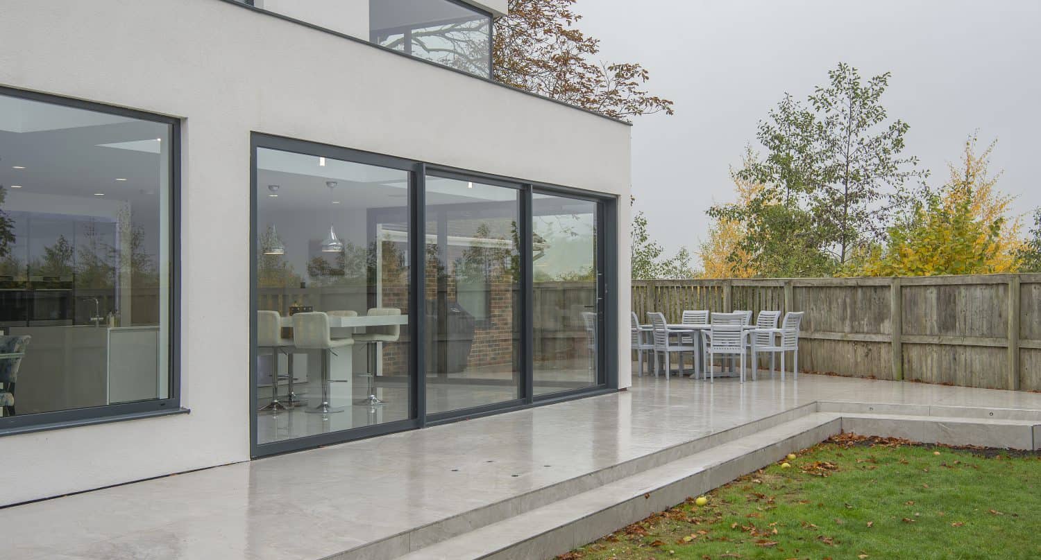 Modern house with large sliding doors opening to a patio with outdoor dining table and chairs, surrounded by a wooden fence and green lawn, with trees in the background and sleek glass roofing overhead.