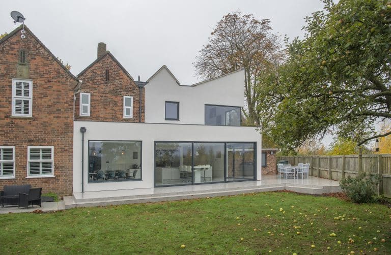 A modern house with a large brick section and a white extension featuring floor-to-ceiling bifold doors, a patio with outdoor seating, and a grassy yard bordered by a wooden fence and trees.