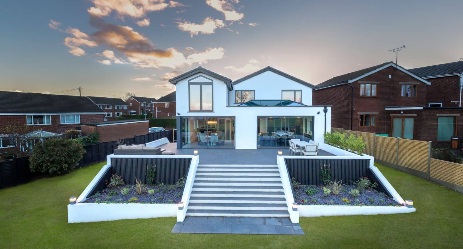 A modern two-story house with large windows, white exterior, and spacious patio featuring elegant sliding doors, set on a well-manicured lawn with raised garden beds and wide steps leading to the entrance; neighboring houses are visible.