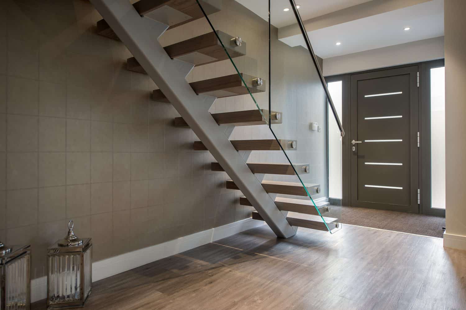 Modern hallway with wooden flooring, a floating wooden staircase with glass railing, taupe walls, decorative lanterns, and a dark front door with horizontal glass panels. Natural light enters from windows beside the door and sleek sliding doors.