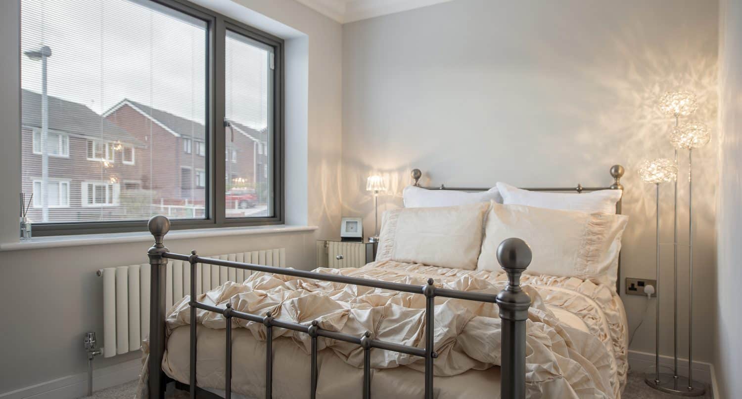 A cozy bedroom with a metal bed frame, white bedding, and pillows sits next to a large window with blinds. Soft lighting and neutral walls enhance the inviting atmosphere, while sliding doors add a modern touch to the space.