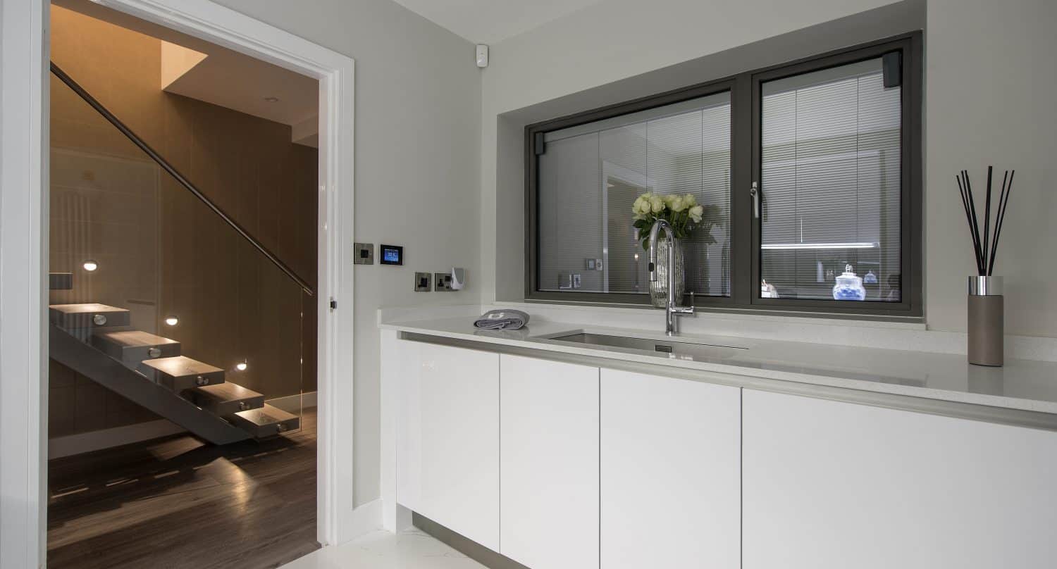 Modern kitchen with white cabinets and a countertop, a large window with blinds, vase of white flowers, and reed diffuser. Doorway opens to stairs with illuminated steps and bespoke glass solutions, set on a stunning marble tile floor.