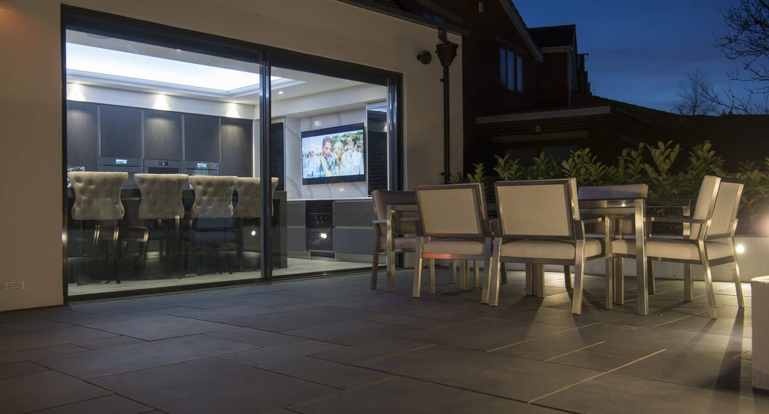 Modern outdoor patio with a dining table and chairs, next to a house featuring sliding doors and large glass panels that reveal a brightly lit kitchen and TV inside, photographed at dusk.