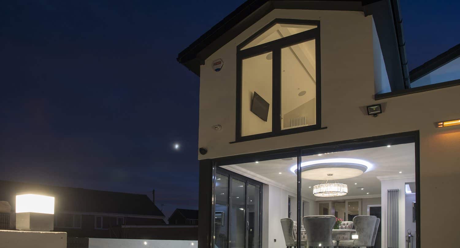 A modern house at night with large glass windows and bespoke glass solutions revealing a well-lit dining area inside. The exterior is illuminated by a garden lamp, and the sky is dark with a visible moon.