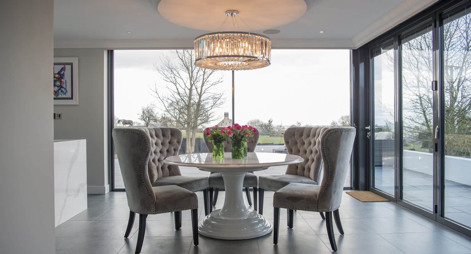 Modern dining room with a round white table, four tufted gray chairs, and a large chandelier above. A bouquet of flowers graces the table, and floor-to-ceiling sliding doors reveal an outdoor view with trees.