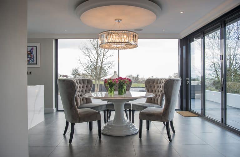Modern dining room with a round white table, four tufted gray chairs, and a large chandelier above. A bouquet of flowers graces the table, and floor-to-ceiling sliding doors reveal an outdoor view with trees.