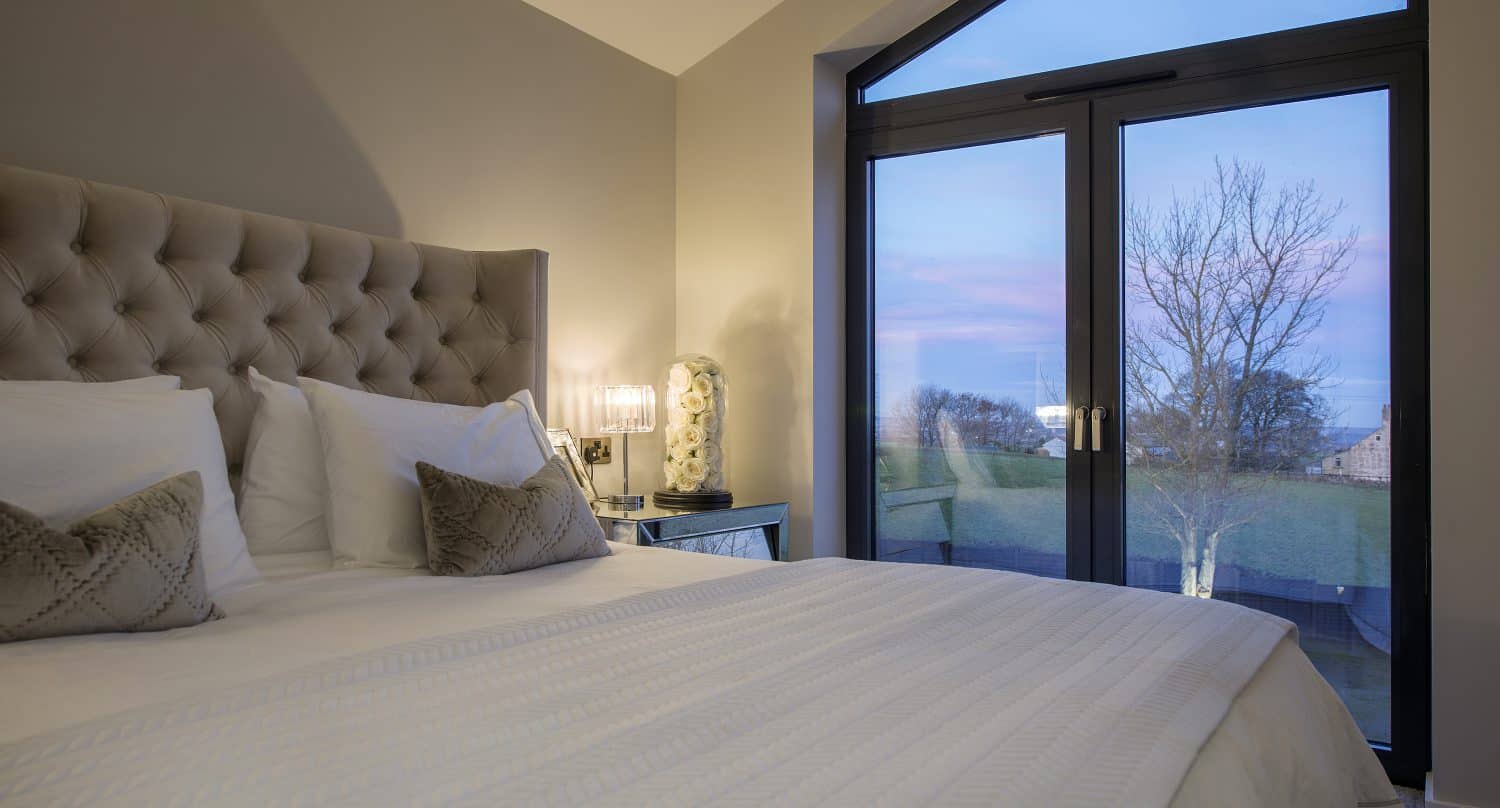 A cozy bedroom with a large bed, tufted headboard, and two decorative pillows sits beside a lamp-lit table. Bespoke glass solutions in the large doors reveal a tree and colorful evening sky just outside.