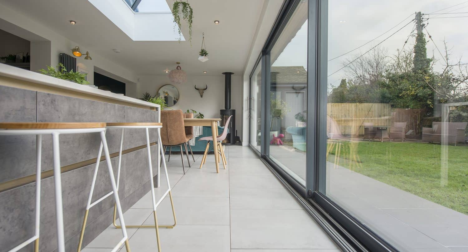 Modern kitchen and dining area with large sliding doors overlooking a green garden. Contemporary bar stools line the counter, while hanging plants and bespoke glass solutions add a fresh touch to the bright, open space.