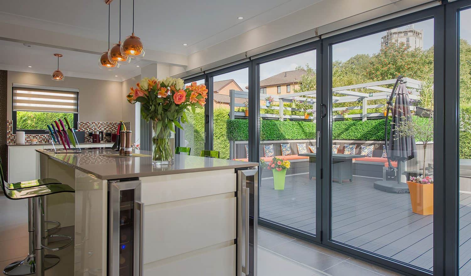 Modern kitchen with a glass island, bar stools, and hanging lights opens to a spacious patio through bespoke glass solutions. The patio features seating, plants, and a pergola beneath glass roofing, with greenery and buildings in the background.