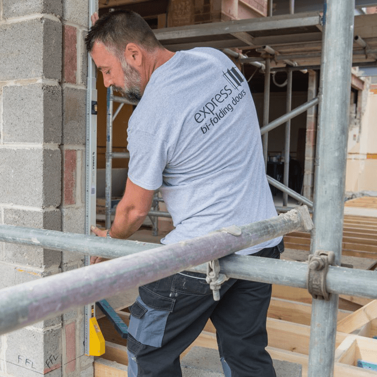 A construction worker in a gray Express Bi-folding Doors t-shirt uses a spirit level to check a brick wall on a building site. Scaffolding and materials surround him, highlighting the precision behind bespoke glass solutions and impressive front doors.
