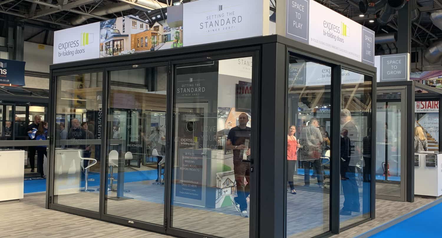 A modern glass showroom display with sliding doors and signage for Express and Standard showcases bespoke glass solutions. People are visible inside and around the booth, which is set on a blue carpet at an indoor exhibition.