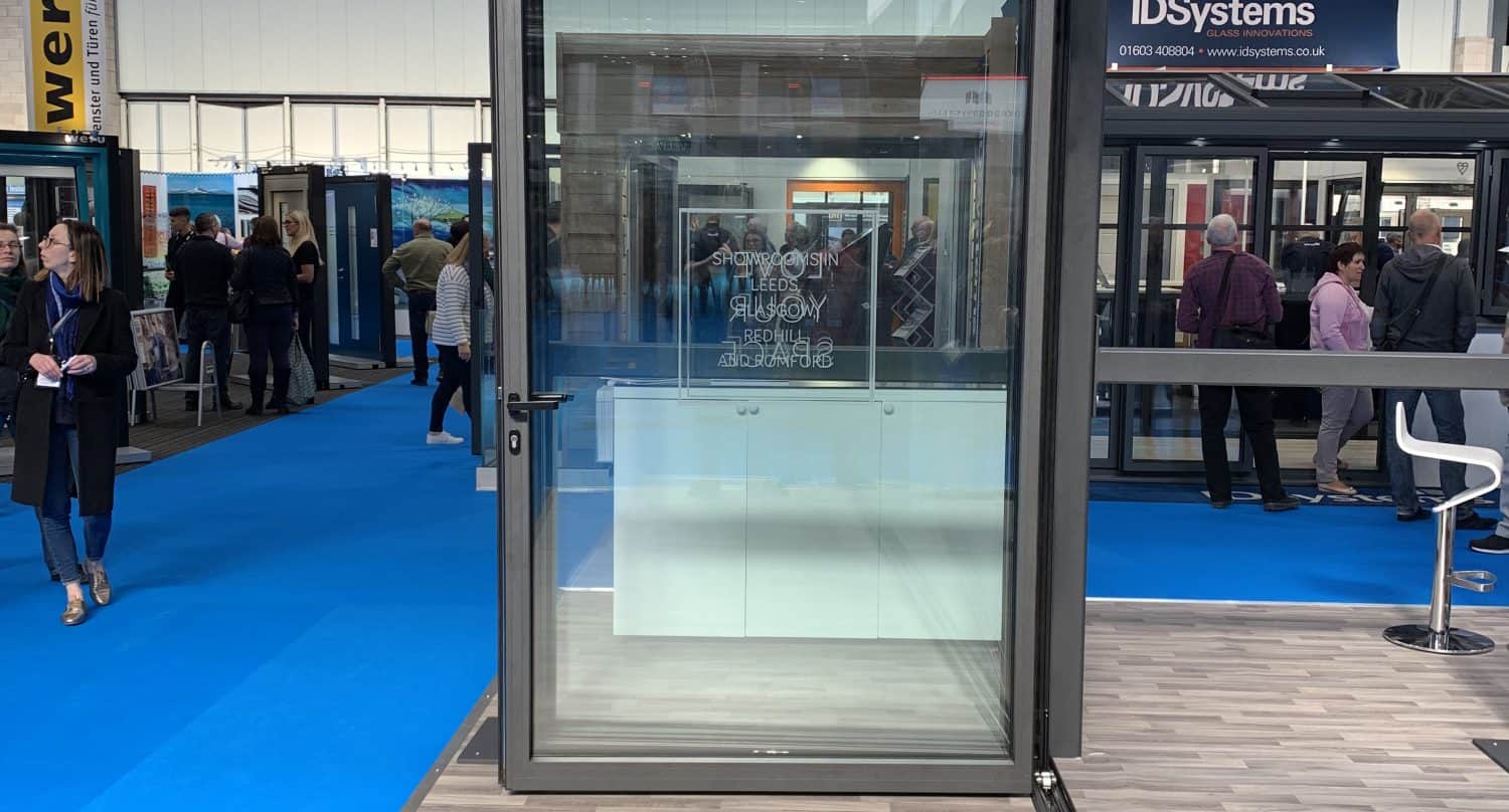 A large glass sliding door with a black frame is displayed in an exhibition hall with people, blue carpet, and various booths in the background. The floor near the door features a wood-patterned design.