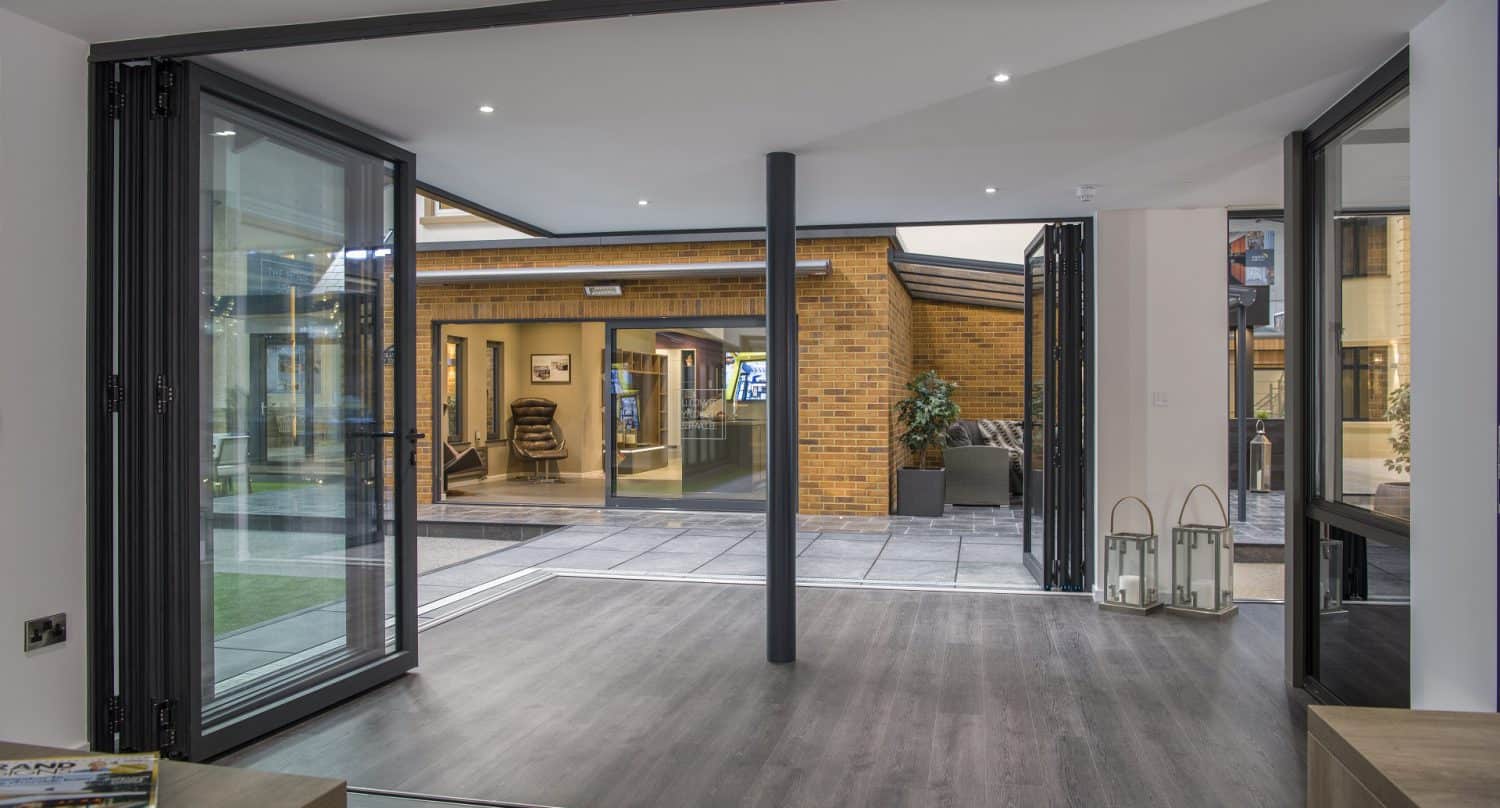 Modern open-plan room with bespoke glass solutions, large glass folding doors opening to a courtyard, featuring brick walls outside, a grey floor indoors, and a few decorative items near the entrance.