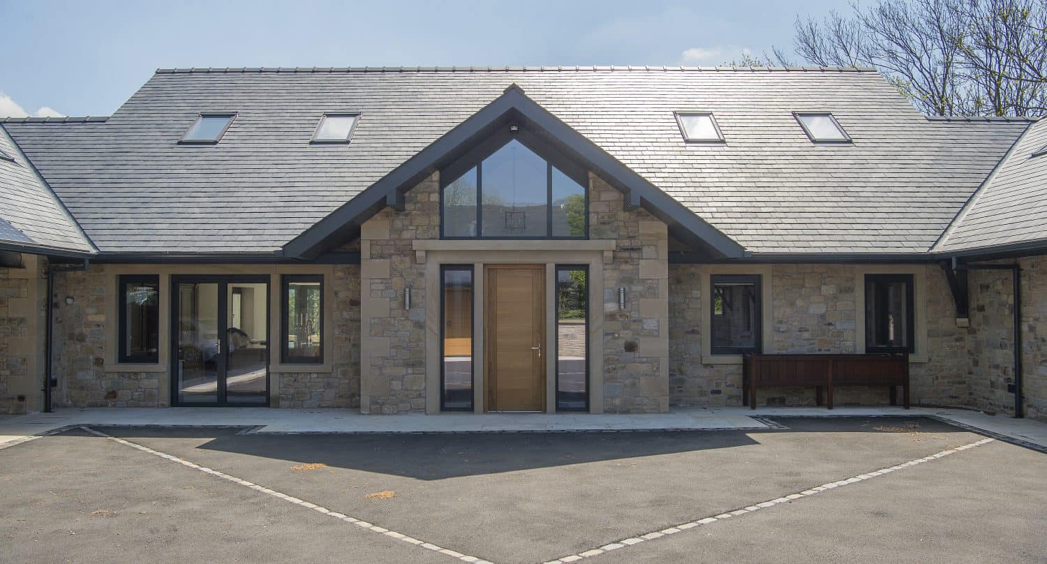 Modern stone house with a large wooden front door, bespoke glass solutions featured in the tall triangular window above, gray tiled roof, and several windows. The house has a paved driveway and is surrounded by trees under a clear blue sky.