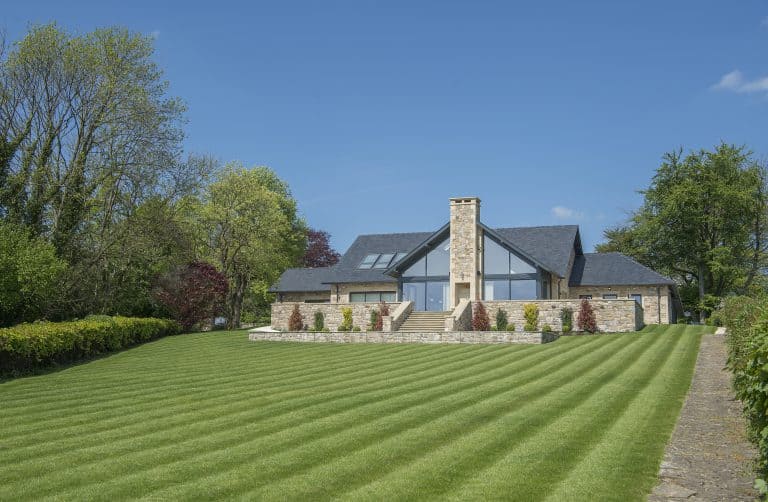 A modern stone house with bespoke glass solutions, including large windows and bifold doors, sits at the top of a wide, neatly-mowed lawn, surrounded by green trees and shrubs under a clear blue sky.