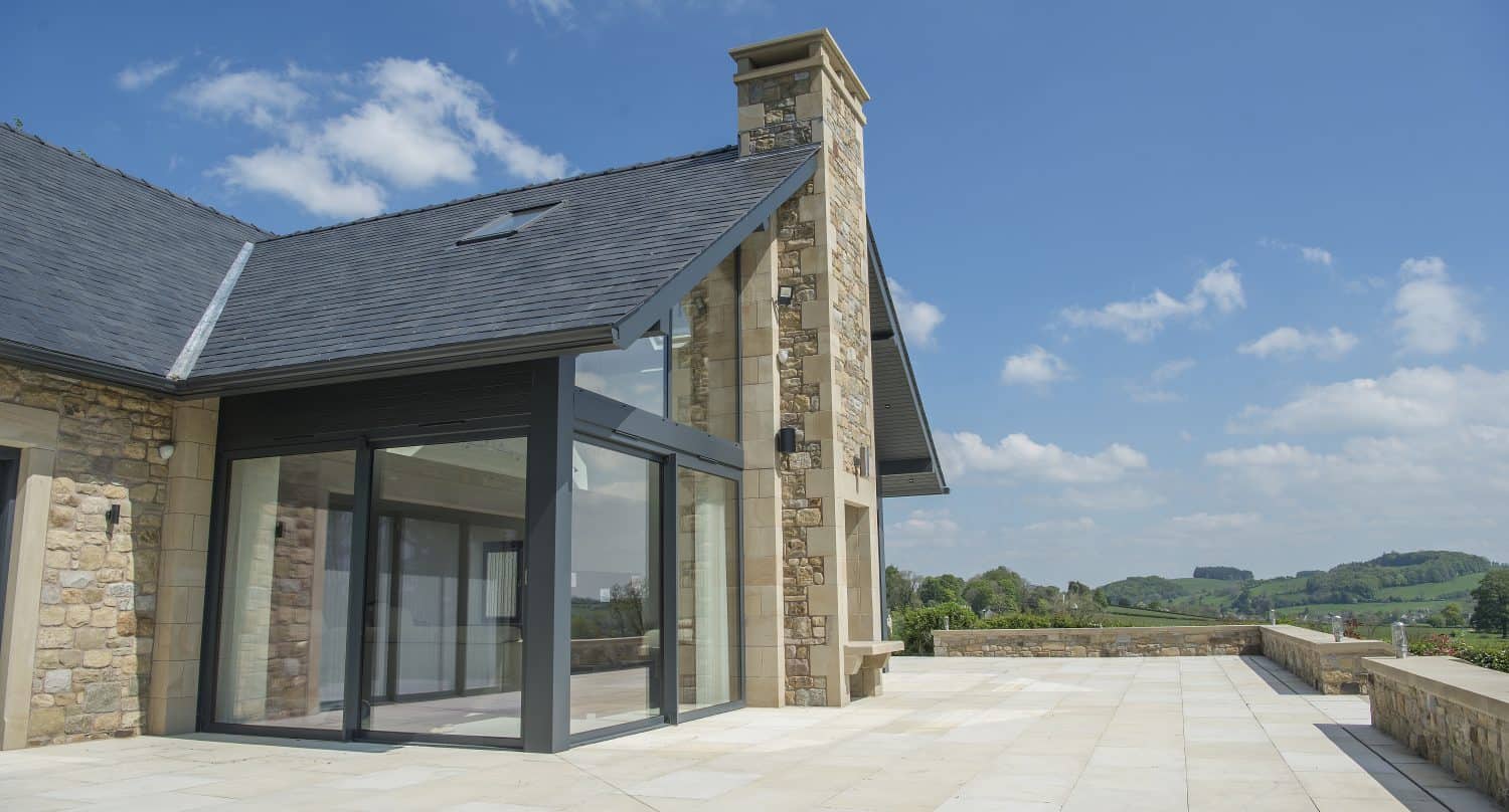 Modern house with stone and glass exterior, large chimney, and spacious tiled patio featuring sliding doors that open to the green countryside under a bright blue sky with scattered clouds.