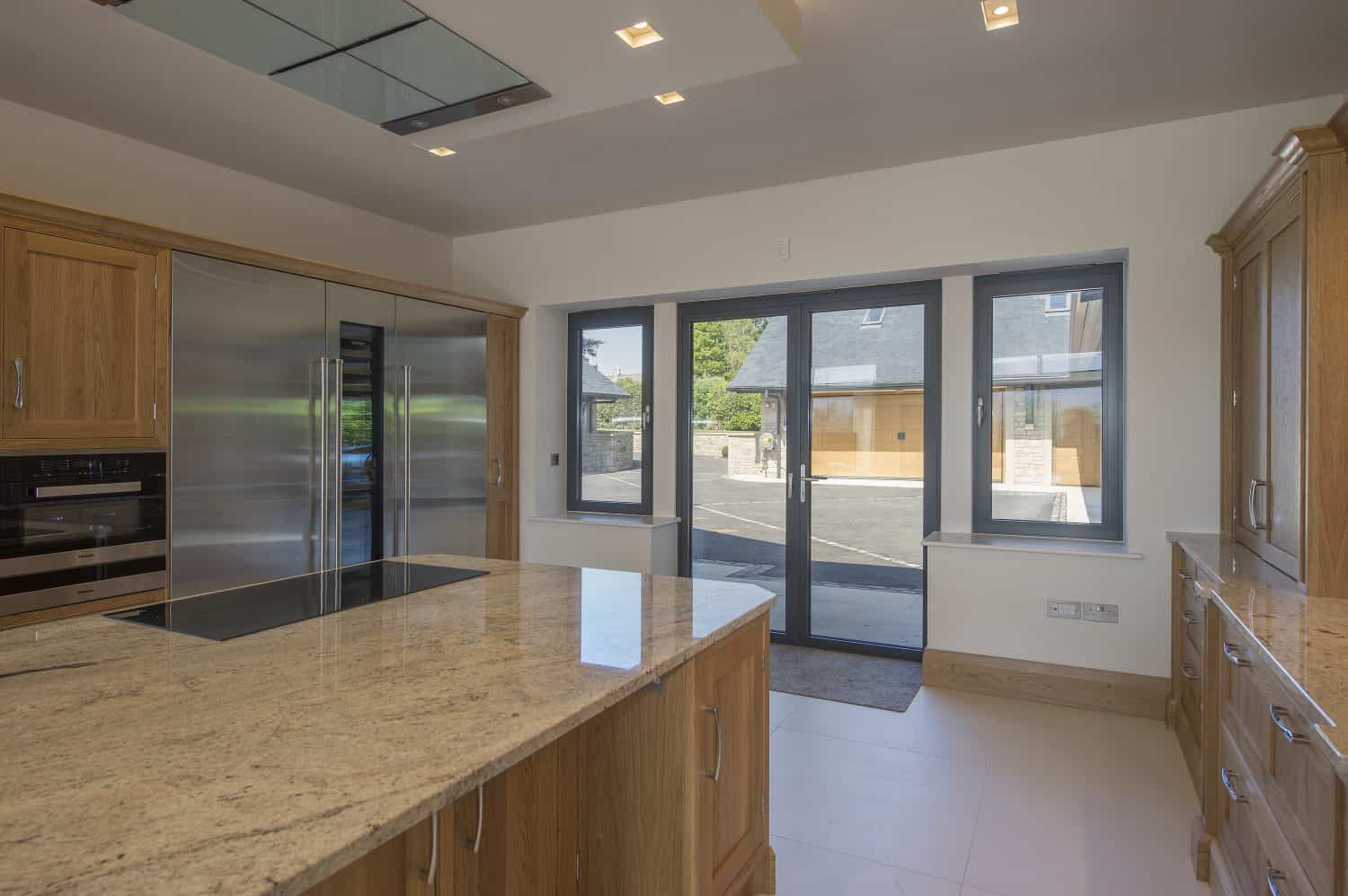 Modern kitchen with wooden cabinets, a large granite island countertop, stainless steel appliances, built-in oven, and expansive bespoke glass solutions including large windows and sliding doors overlooking buildings and surrounding greenery.