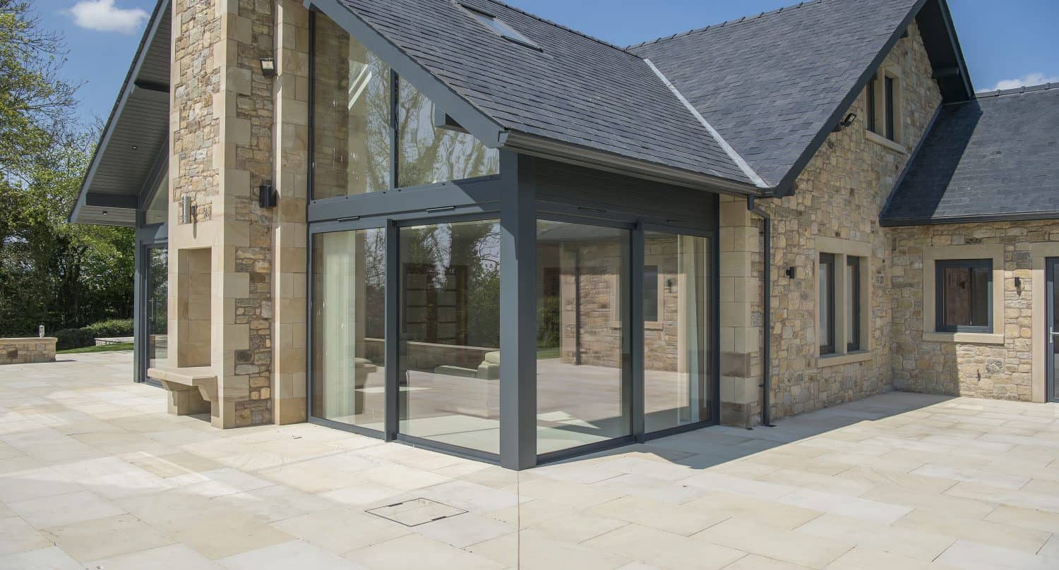 Modern stone house with large glass windows and bifold doors, a gray tiled roof, and a spacious paved patio area under a clear blue sky.