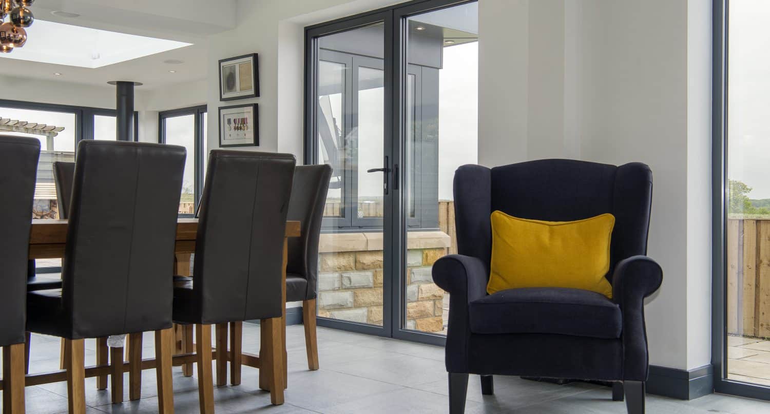 Modern dining area with a wooden table and black leather chairs. A dark blue armchair with a yellow cushion sits near large windows featuring bespoke glass solutions, providing views of the patio.